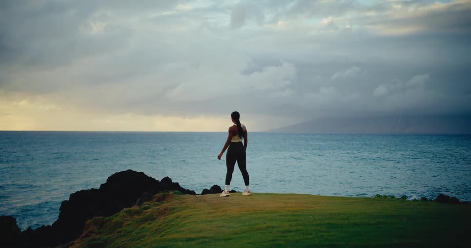 An athletic woman jogs in slow motion across a grassy clifftop before stopping to take in the scenic ocean view