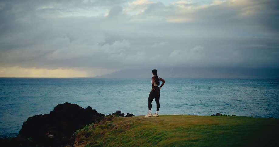 An athletic woman jogs along a scenic, grassy coastline at dusk and stops to admire the beautiful ocean view