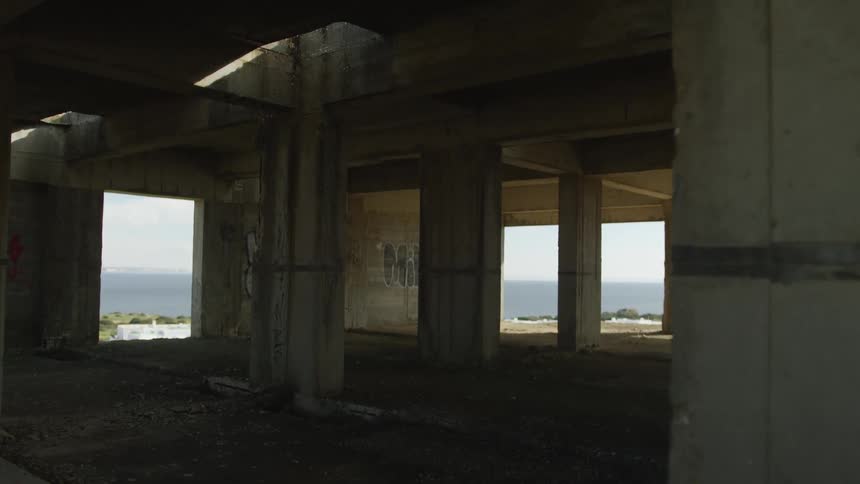 Slow zoom out from inside a dark, derelict concrete building with large open walls framing a bright ocean view