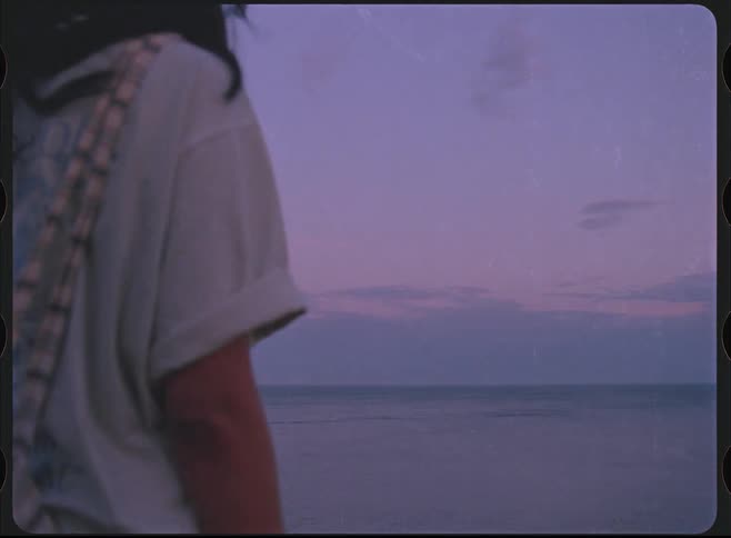A person with long dark hair stands looking out at the purple ocean horizon at sunset in a dreamy retro-style shot