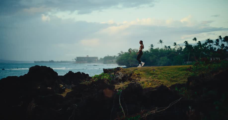 A woman jogs along a scenic tropical cliffside path by the ocean at dusk, stopping to admire the view