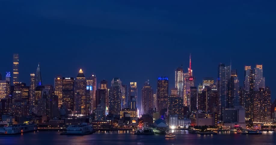 The iconic Midtown Manhattan skyline glitters with colorful lights against the deep blue night sky as boats pass by on the river