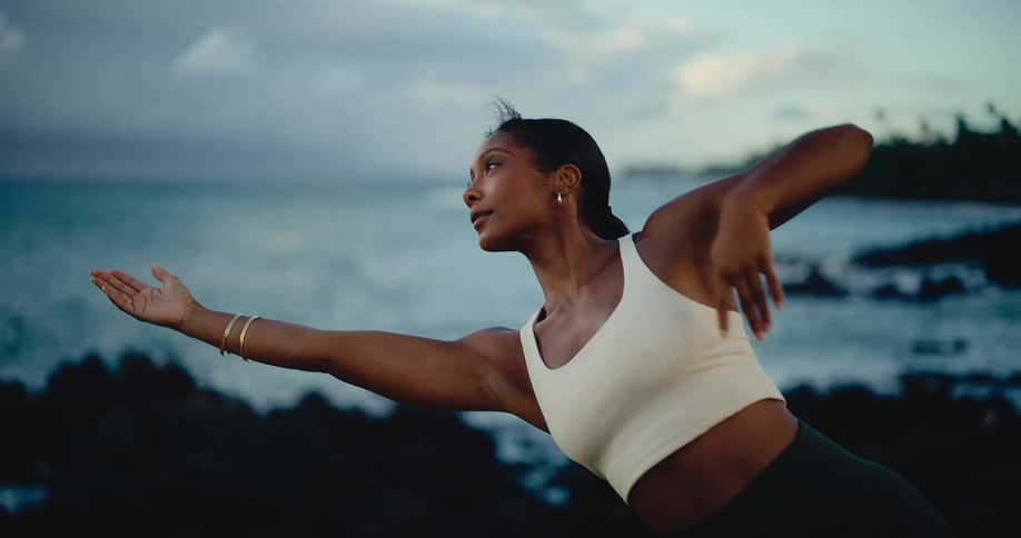 A beautiful woman gracefully flows through yoga poses on a green cliff overlooking the ocean at sunset