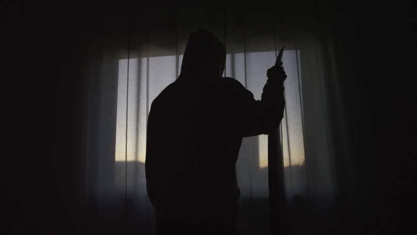 The menacing silhouette of a hooded killer stands with a knife in a dark room