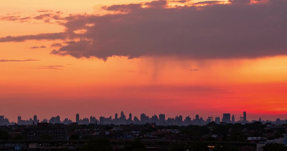 A dramatic timelapse shows a fiery sunset over a city skyline with distant rain showers and passing subway trains