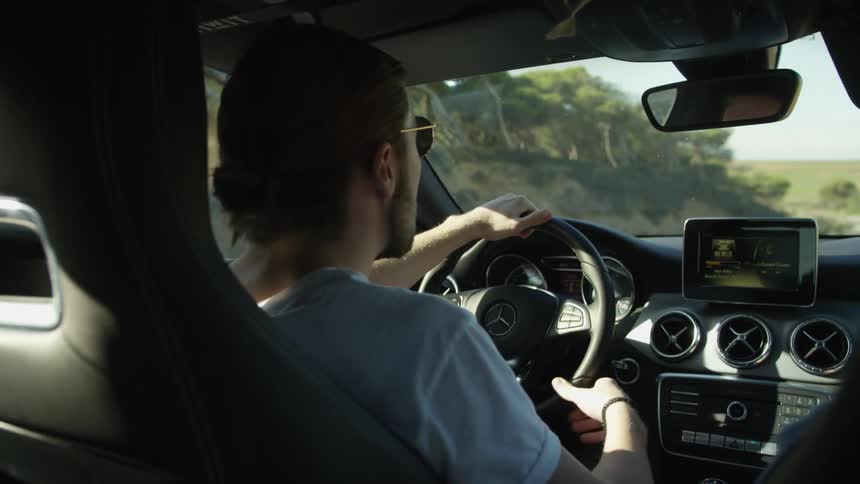 An over-the-shoulder shot of a young man with long hair driving a luxury car along a scenic country road on a sunny day