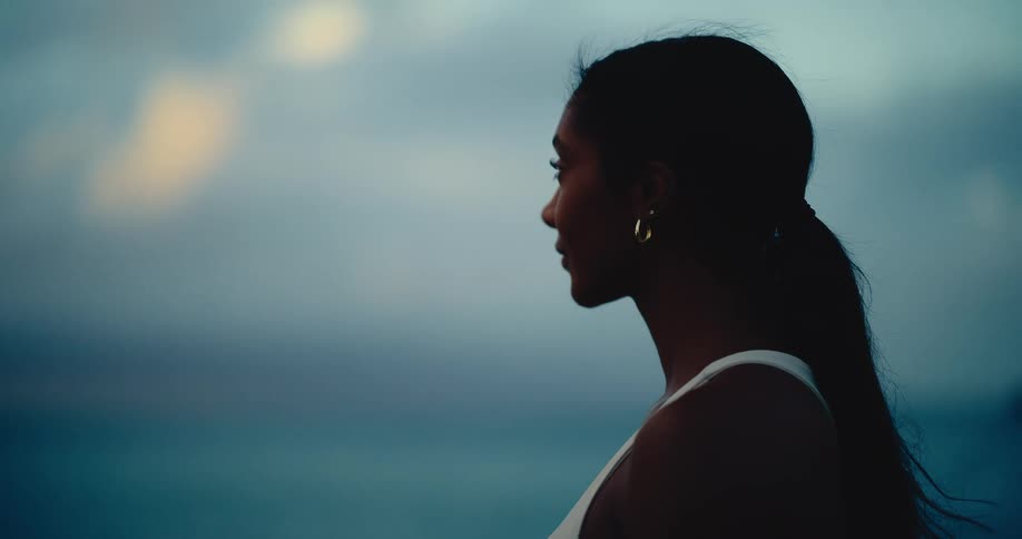 A silhouetted woman in profile looks out peacefully at the ocean during a moody twilight