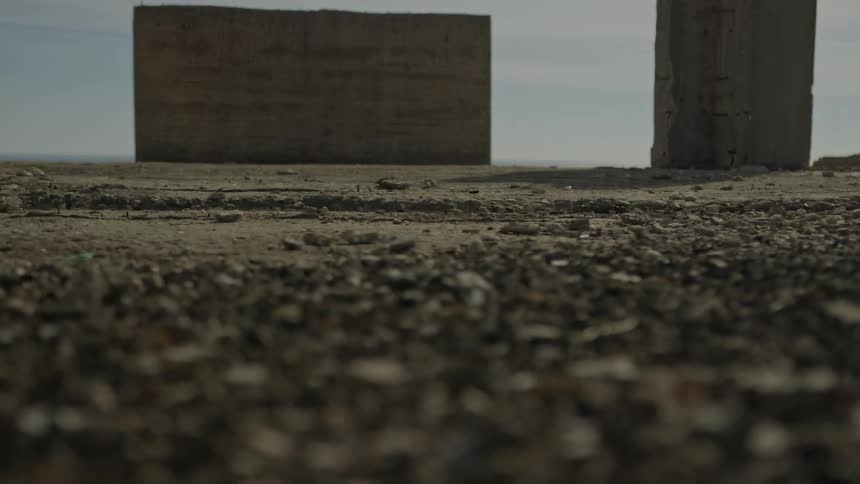 A slow rack focus from a dusty, gravel-covered ground reveals abandoned concrete structures under a hazy sky