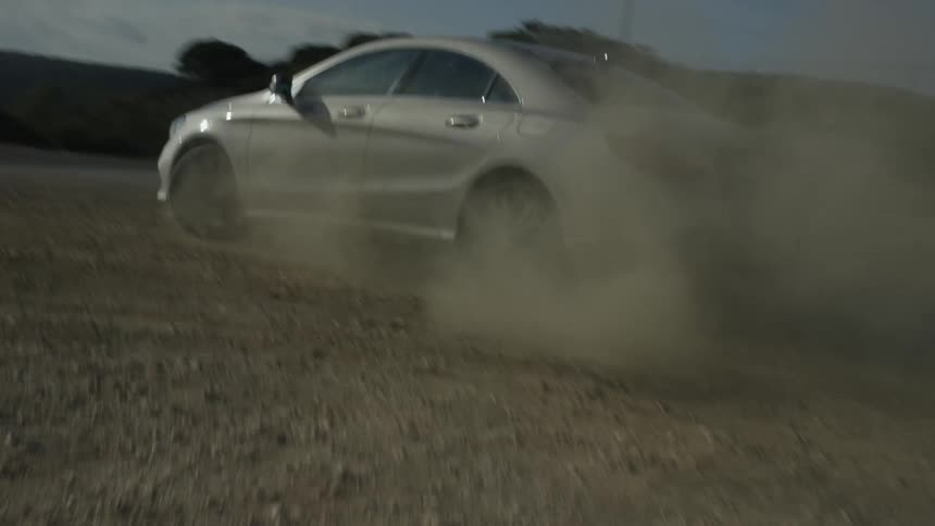 A silver luxury sedan kicks up a massive cloud of dust as it speeds along a rugged gravel road in slow motion