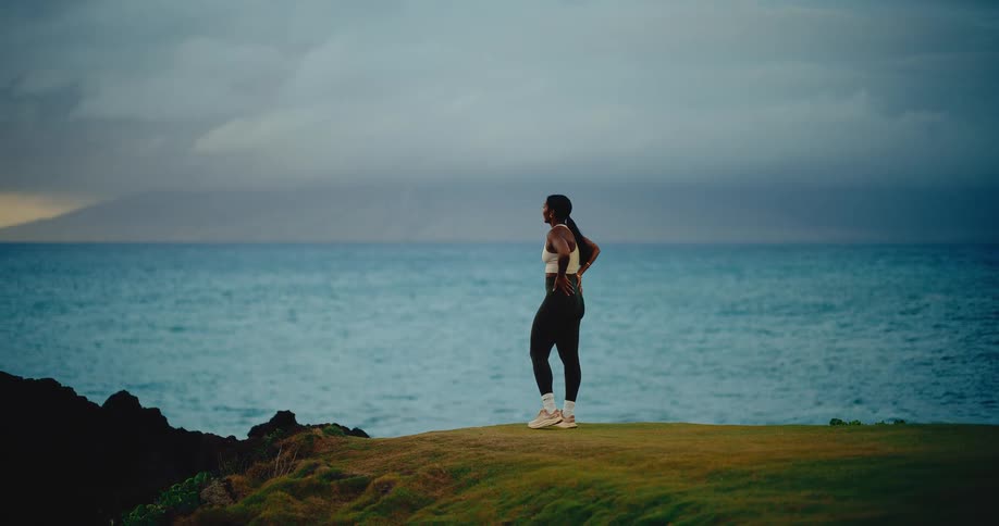 A fit woman runs in slow motion along a grassy cliffside before stopping to admire the ocean view at dusk