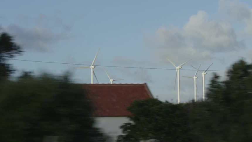 A tracking shot from a moving vehicle captures a wind farm with spinning turbines on a lush green hillside