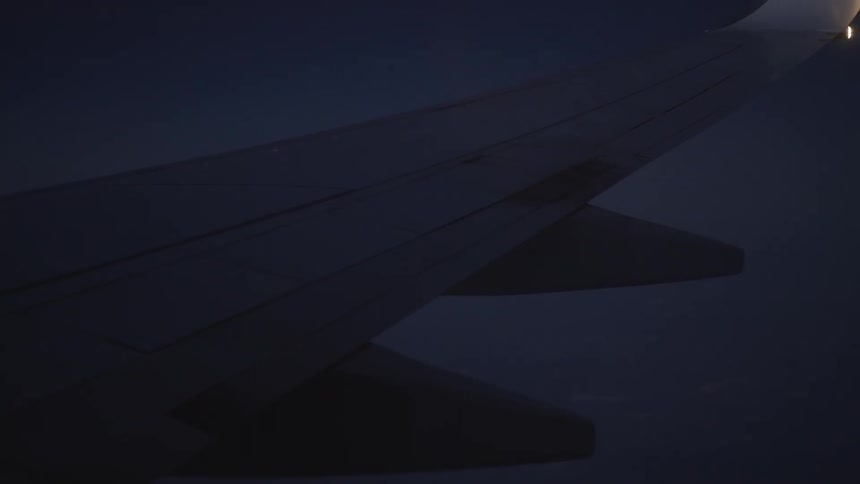 A close-up view from an airplane window of the wing flying through the dark night sky with flashing navigation lights