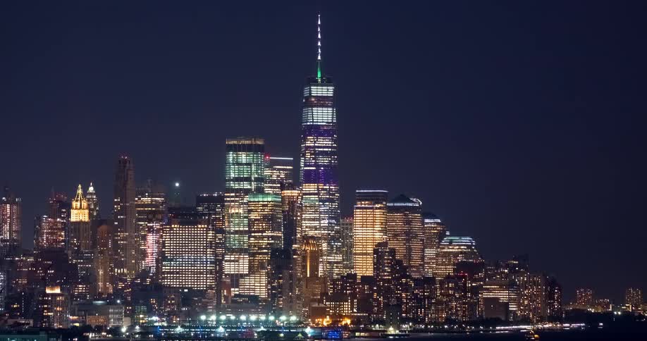 A time-lapse view shows the brightly lit lower Manhattan skyline and One World Trade Center against a dark night sky