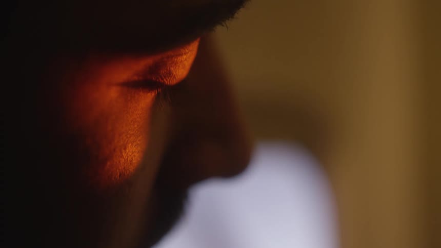 A moody, intimate close-up of a man's eye reflecting a warm, orange light as he gazes downward in a dark room