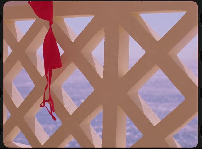 A red bikini top hangs on a white patterned balcony railing with a shimmering ocean in the background, captured in a vintage style