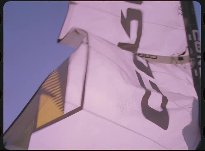 A shadowy figure holds a large kitesurfing kite as it billows in the wind, with the camera tilting up to the bright sun in a purple sky