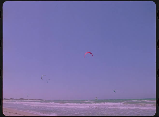 Lo-fi, vintage-style footage captures a serene scene of kitesurfing at the beach under a clear sky