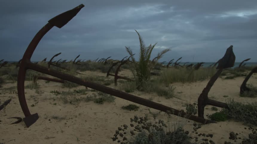 A desolate landscape of rusty, abandoned anchors scattered across a sandy beach under a dark and cloudy sky