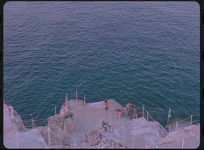 A lone man jumps in slow motion from a rocky cliffside pier into the ocean in a vintage film-style shot