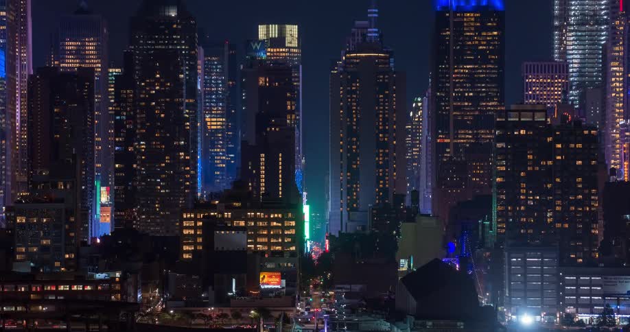 A vibrant nighttime timelapse captures the bustling energy of a city street canyon, flanked by towering, illuminated skyscrapers