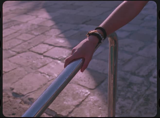 A woman in a swimsuit approaches a metal railing by a stone poolside and places her hand on it in a vintage-style clip