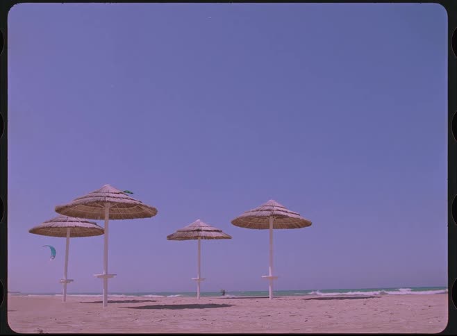 Vintage 8mm film style footage of straw umbrellas on a sandy beach with people kitesurfing in the distance