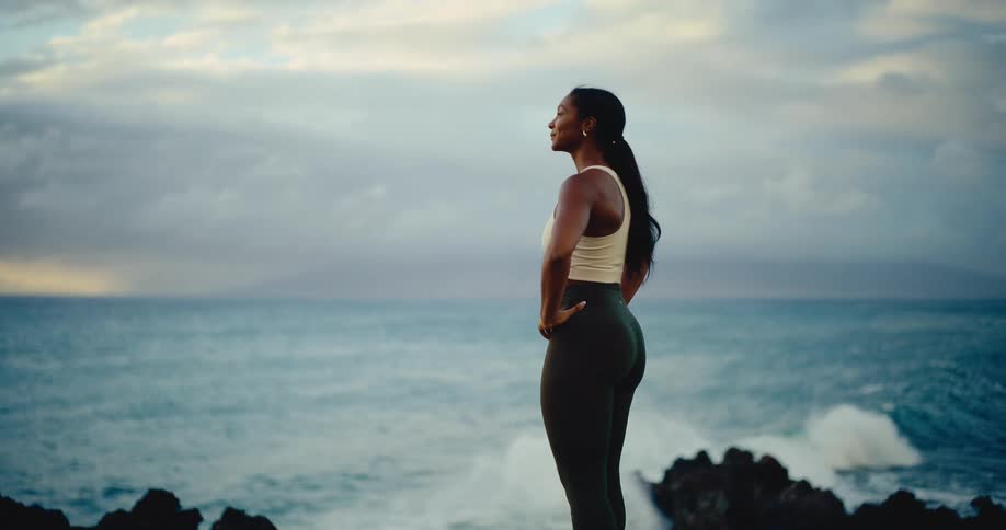 A fit athletic woman runs along a beautiful tropical coastline and pauses to admire the powerful ocean waves crashing at sunset
