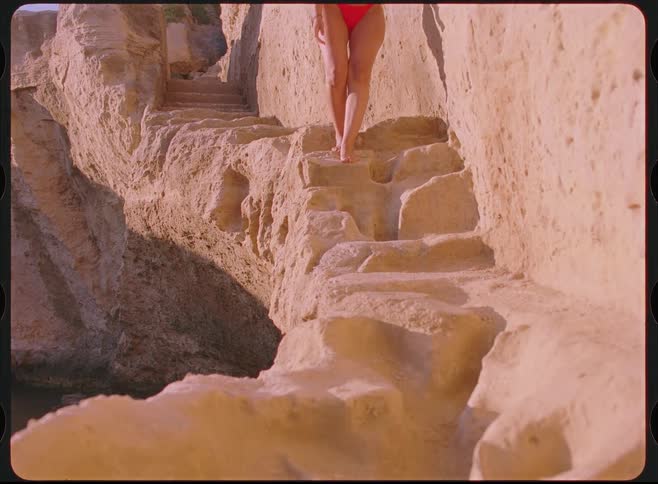 Vintage film footage shows a woman in a red bikini walking down stairs carved into a seaside rock cliff