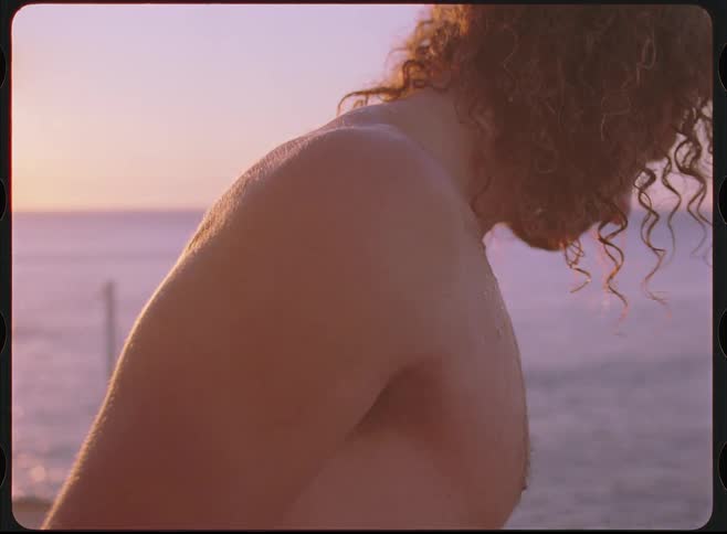 A shirtless man with long curly hair shakes water off in slow motion by the ocean during a golden sunset