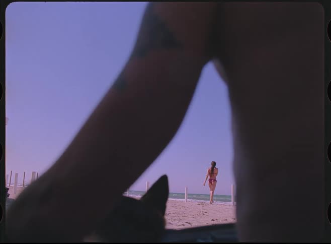 A nostalgic, vintage-style clip shows a woman in a bikini walking towards the ocean on a sunny day, filmed from a low point of view on the sand