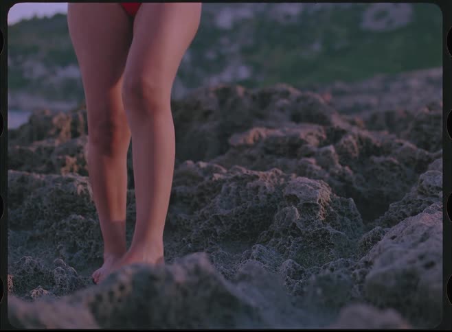 A woman in a red bikini carefully walks barefoot across a rugged, rocky shoreline in slow motion, shot with a vintage film effect