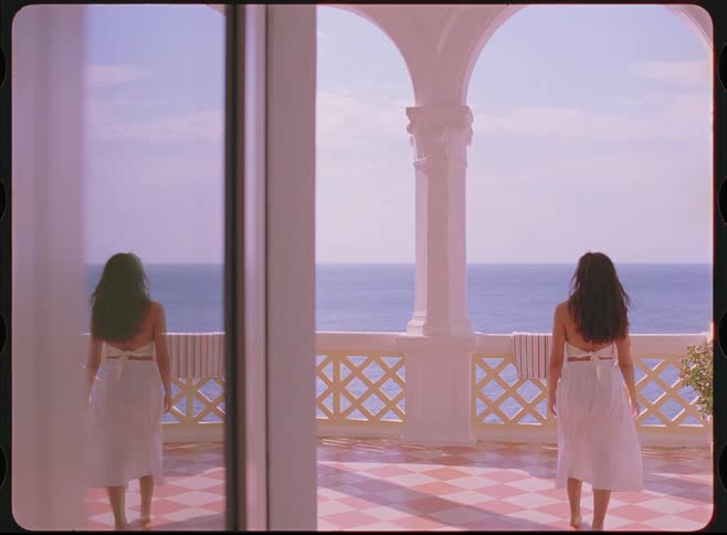 A woman in a flowing white dress walks out onto a sunlit, ornate balcony to admire the tranquil ocean view, captured with a beautiful vintage film aesthetic