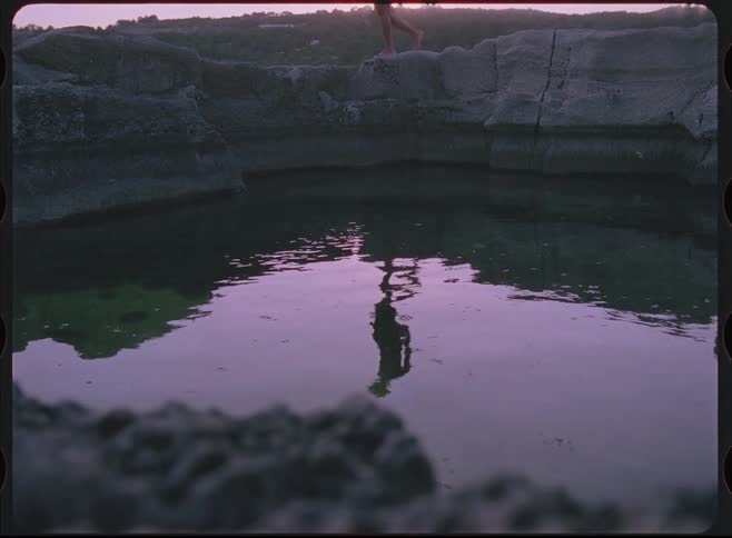 A vintage, dreamlike shot shows the reflection of a person walking alongside a still, purple-hued rock pool at twilight