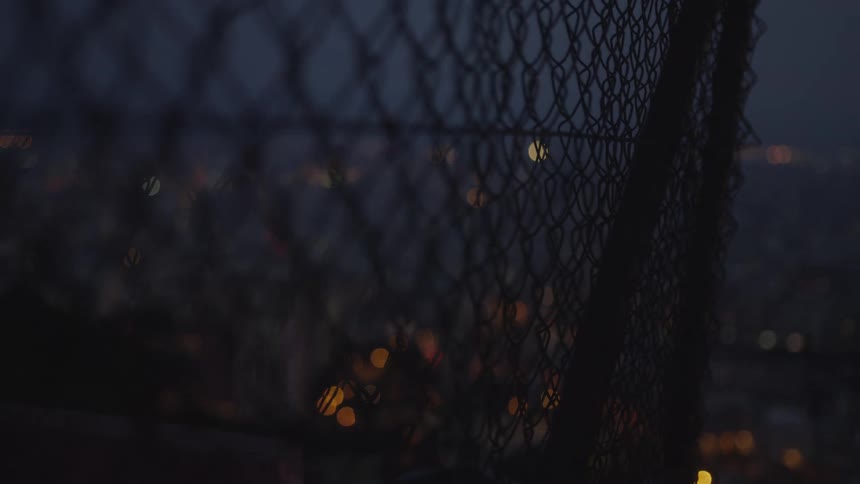 A moody rack focus from a chain-link fence to the bokeh lights of the city skyline at dusk