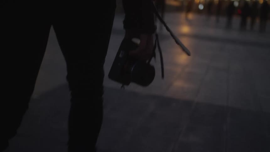A silhouetted photographer walks through a city at dusk, holding a camera in slow motion with glowing streetlights in the background