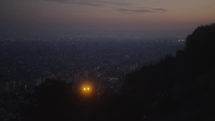 A pair of glowing lights peer out from a dark hillside overlooking the vast, illuminated cityscape at twilight