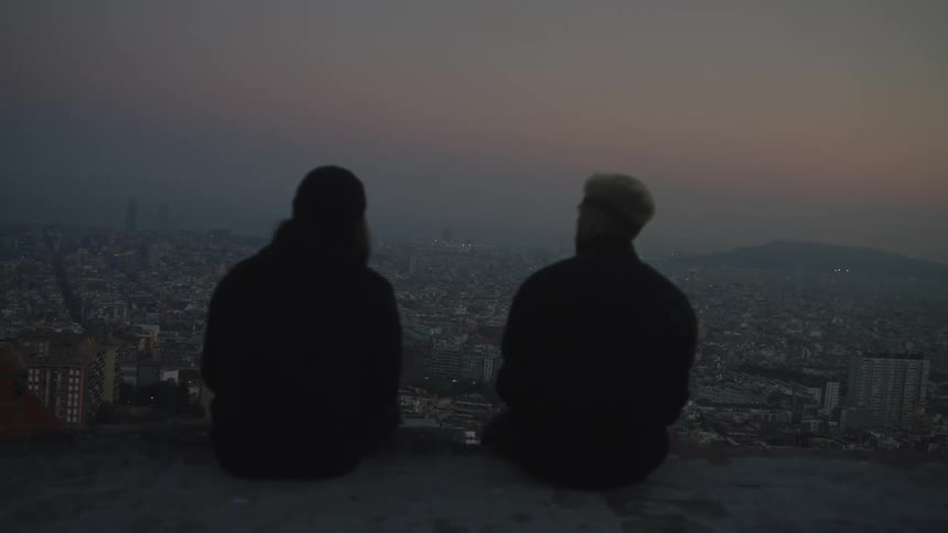 Two friends sit together on a high overlook, quietly watching the sprawling cityscape at twilight