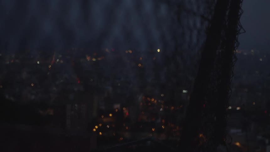 A dramatic focus pull reveals a sprawling cityscape with glowing bokeh lights at night through a chain-link fence