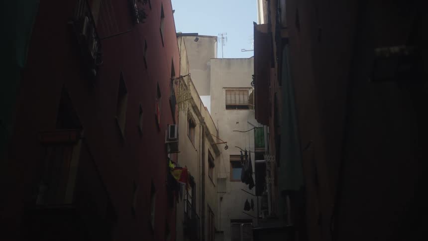 A static, low-angle shot looks up through a dark, narrow alleyway between old apartment buildings in Spain
