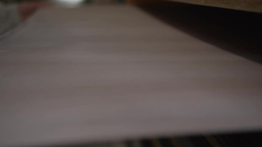 A close-up of a factory worker's hand carefully applying veneer edge banding to a wooden panel using machinery
