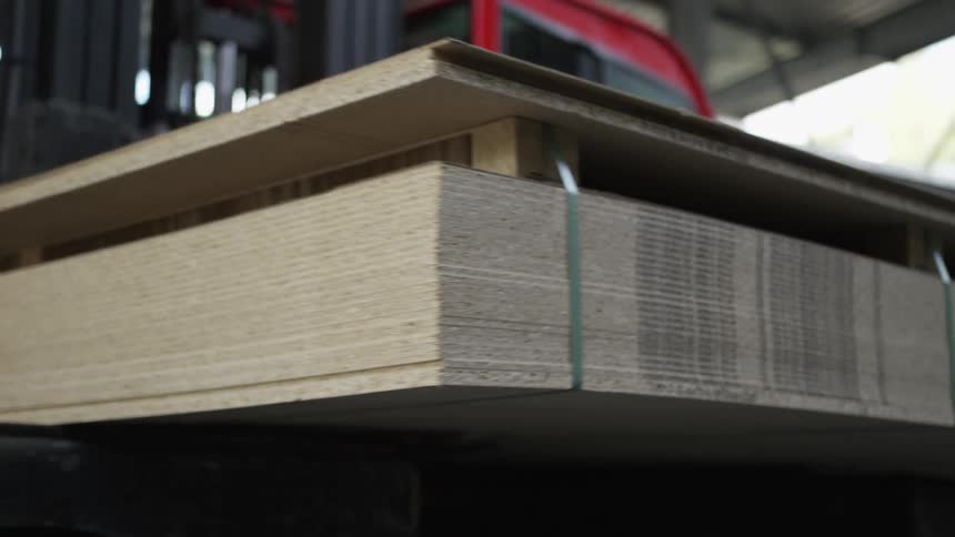 A forklift carefully lifts a pallet of stacked particleboard sheets inside a large industrial warehouse