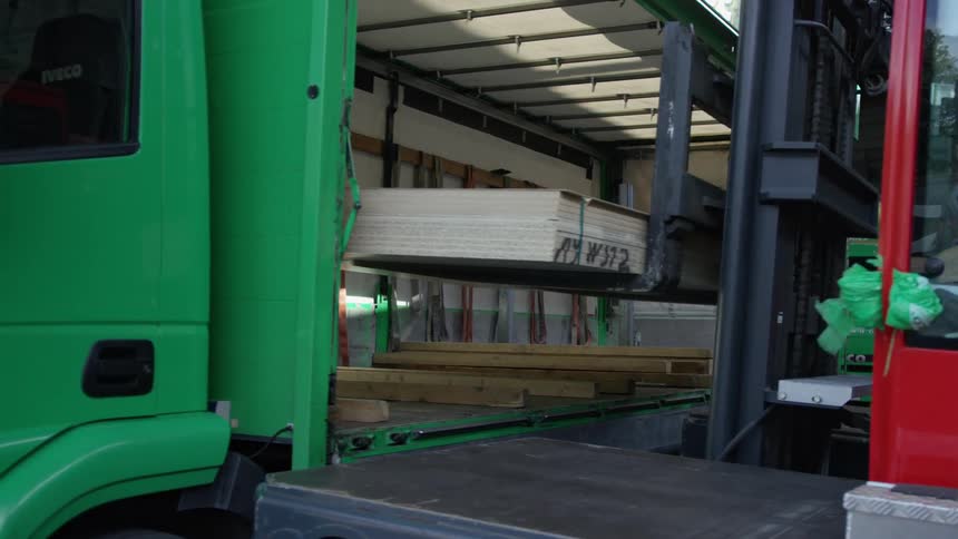 A forklift carefully unloads a large stack of wood panels from the cargo bed of a green truck