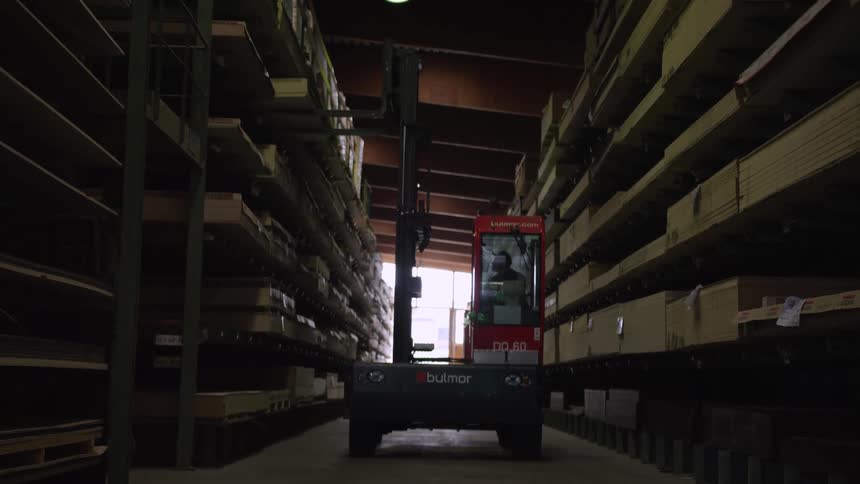 A red forklift lowers its forks while parked in the aisle of a large, dimly lit lumber warehouse