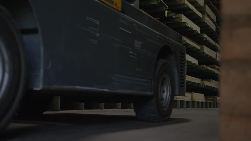 A forklift slowly drives away from the camera in a large industrial warehouse with shelves of lumber