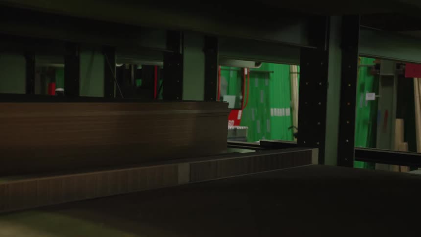 A red sideloader forklift drives through the aisles of a large, dimly lit lumber and wood panel warehouse