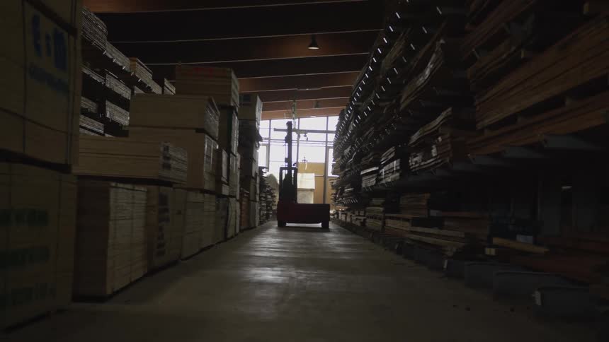 A stationary red forklift sits at the end of a long, dimly lit aisle in a massive lumber warehouse