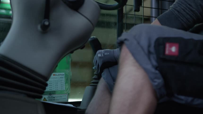 A forklift operator wearing gloves maneuvers the vehicle using the steering wheel and joystick inside a warehouse