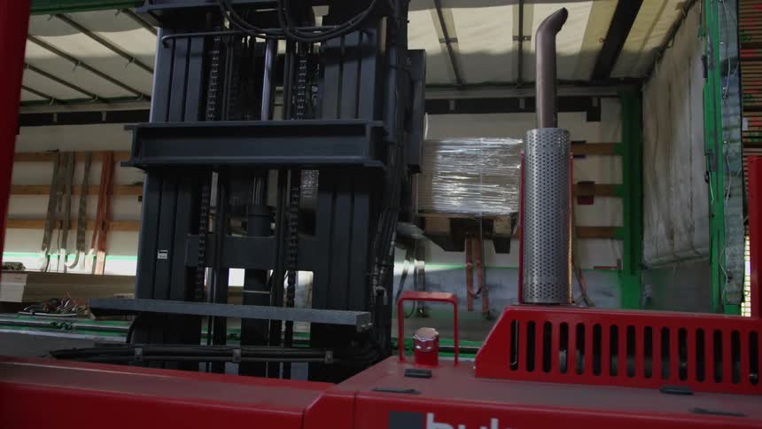 A large red forklift carefully reverses out of a truck trailer, pulling a shrink-wrapped pallet of goods in a warehouse
