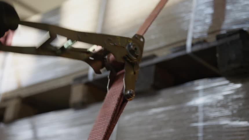 A worker wearing gloves tightens a red ratchet strap in slow motion to secure cargo in a warehouse