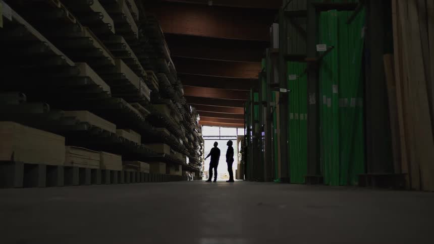 Two silhouetted workers discuss inventory while standing in a long, dark aisle of a lumber warehouse filled with shelves of building materials